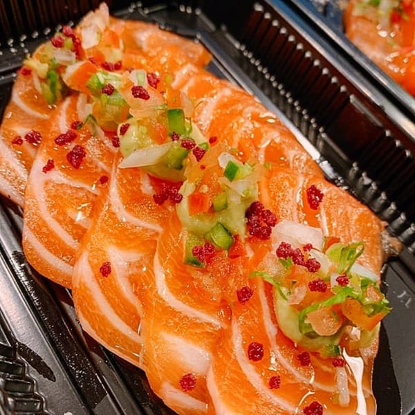 Close-up of salmon sashimi topped with diced vegetables and red sprinkles in a black tray.