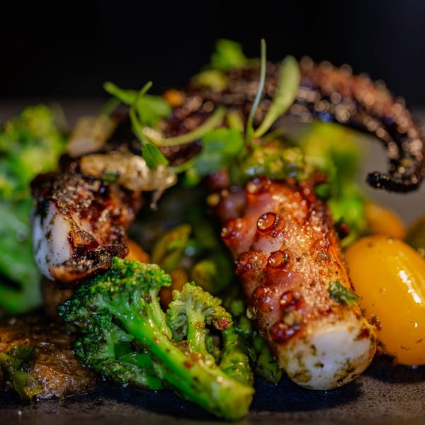Grilled octopus with charred broccoli, yellow tomatoes, and fresh herbs on a dark plate.