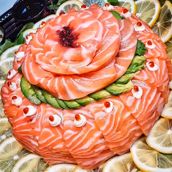 A salmon sashimi rose with avocado, roe, and lemon slices, presented on a dark platter.