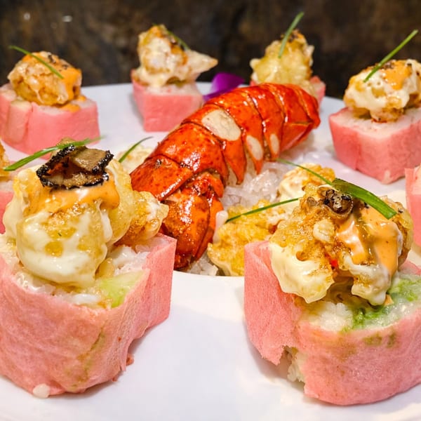 Lobster tail on ice surrounded by pink-wrapped sushi rolls topped with sauce and chives.