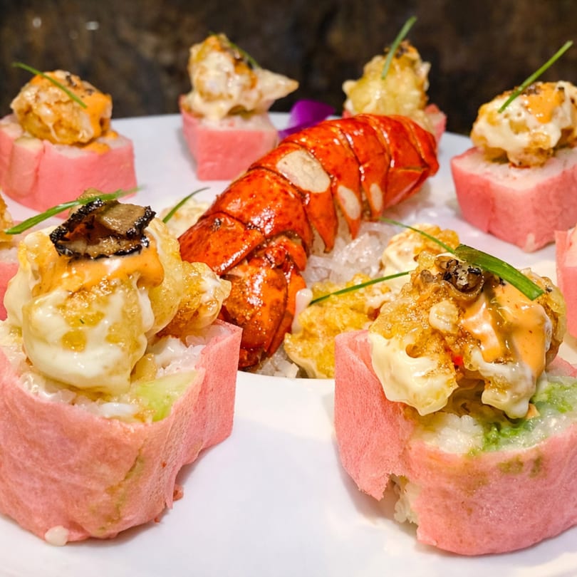 Lobster tail on ice surrounded by pink-wrapped sushi rolls topped with sauce and chives.