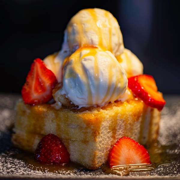 Honey toast dessert with vanilla ice cream, caramel drizzle, and fresh strawberries on a dark plate.