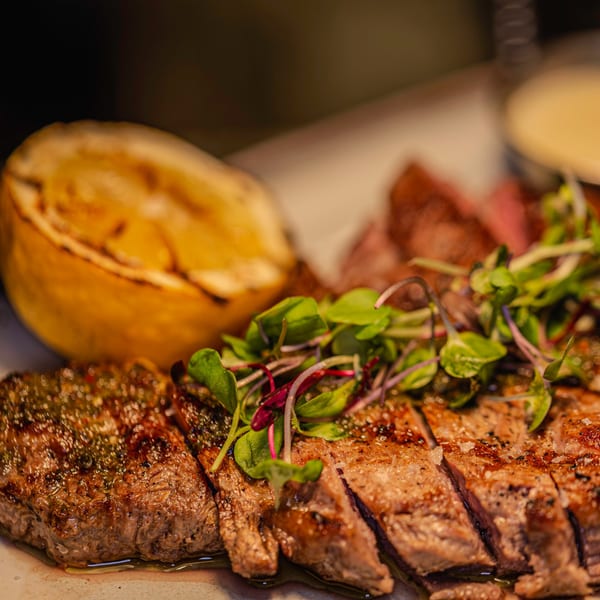 Sliced grilled steak with green herbs and a charred lemon half on a white plate.