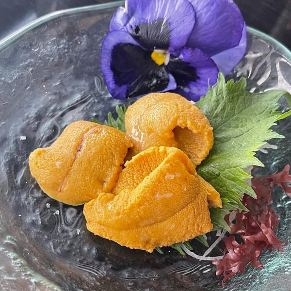 Three pieces of bright orange uni (sea urchin) on a glass plate with green shiso leaf and a purple flower.